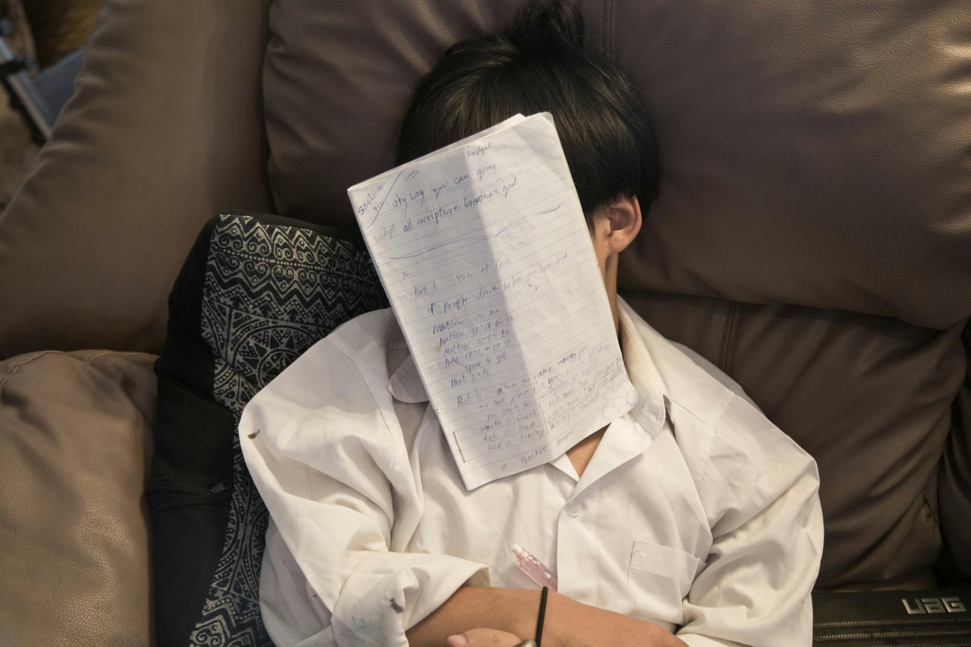 A person in white shirt naps on a couch with a notebook on face, depicting exhaustion.