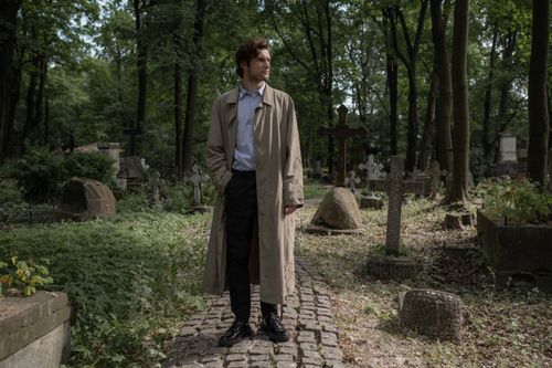 Free A man in a trench coat stands in a serene cemetery setting surrounded by lush trees. Stock Photo