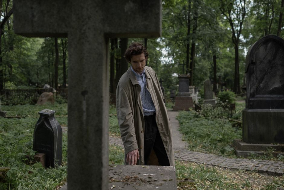 Caucasian man in trench coat mourns at a rustic cemetery during the day.