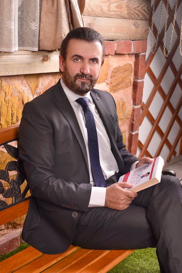 A Man In A Suit Holding A Book While Sitting On A Bench