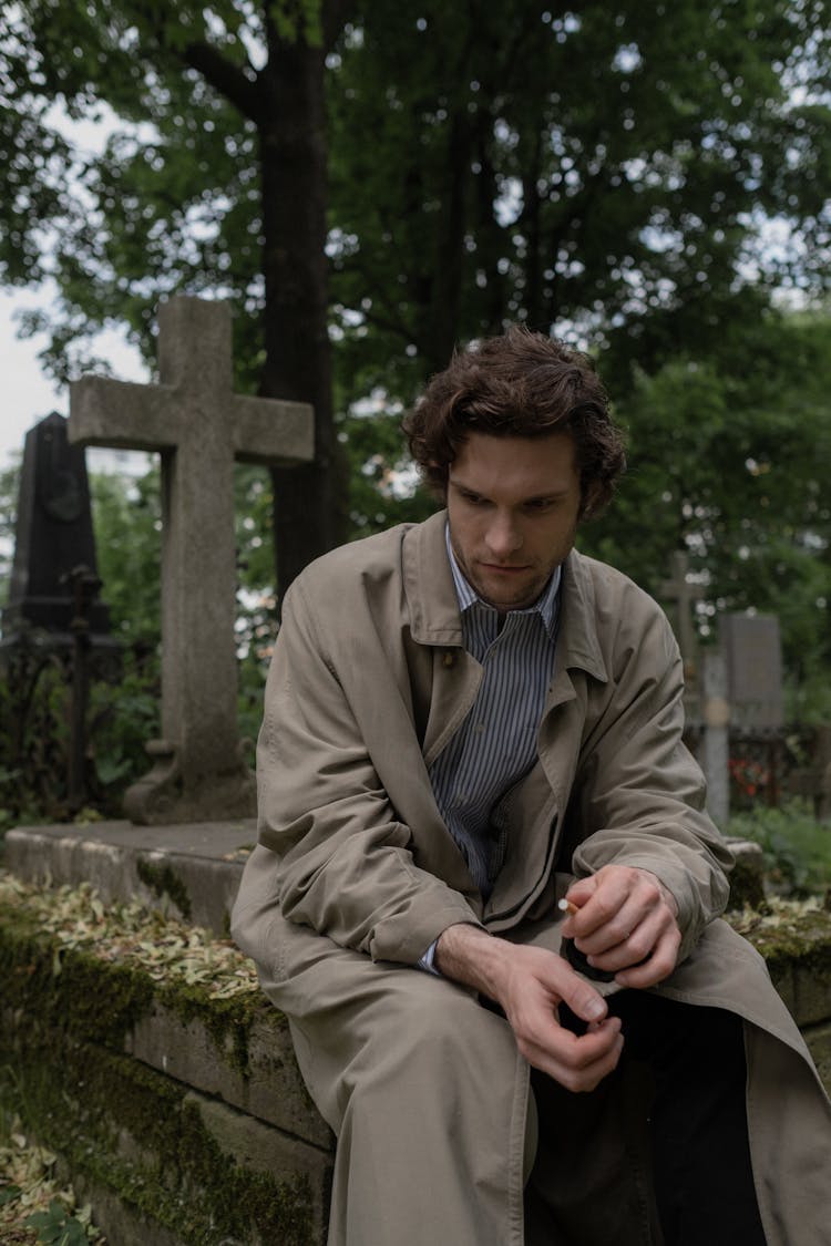 A Man In A Trench Coat Sitting On A Grave