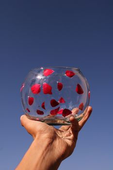 A clear fish bowl filled with red petals held up against a bright blue sky.