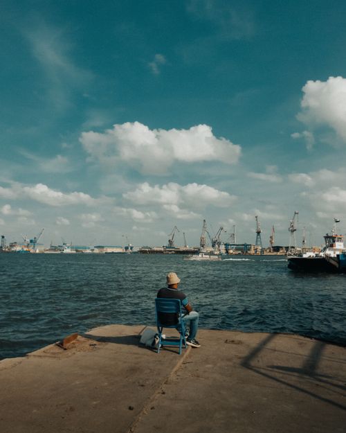 Free A man sitting on a chair by the water at Port Said harbor, Egypt, overlooking boats and cranes. Stock Photo