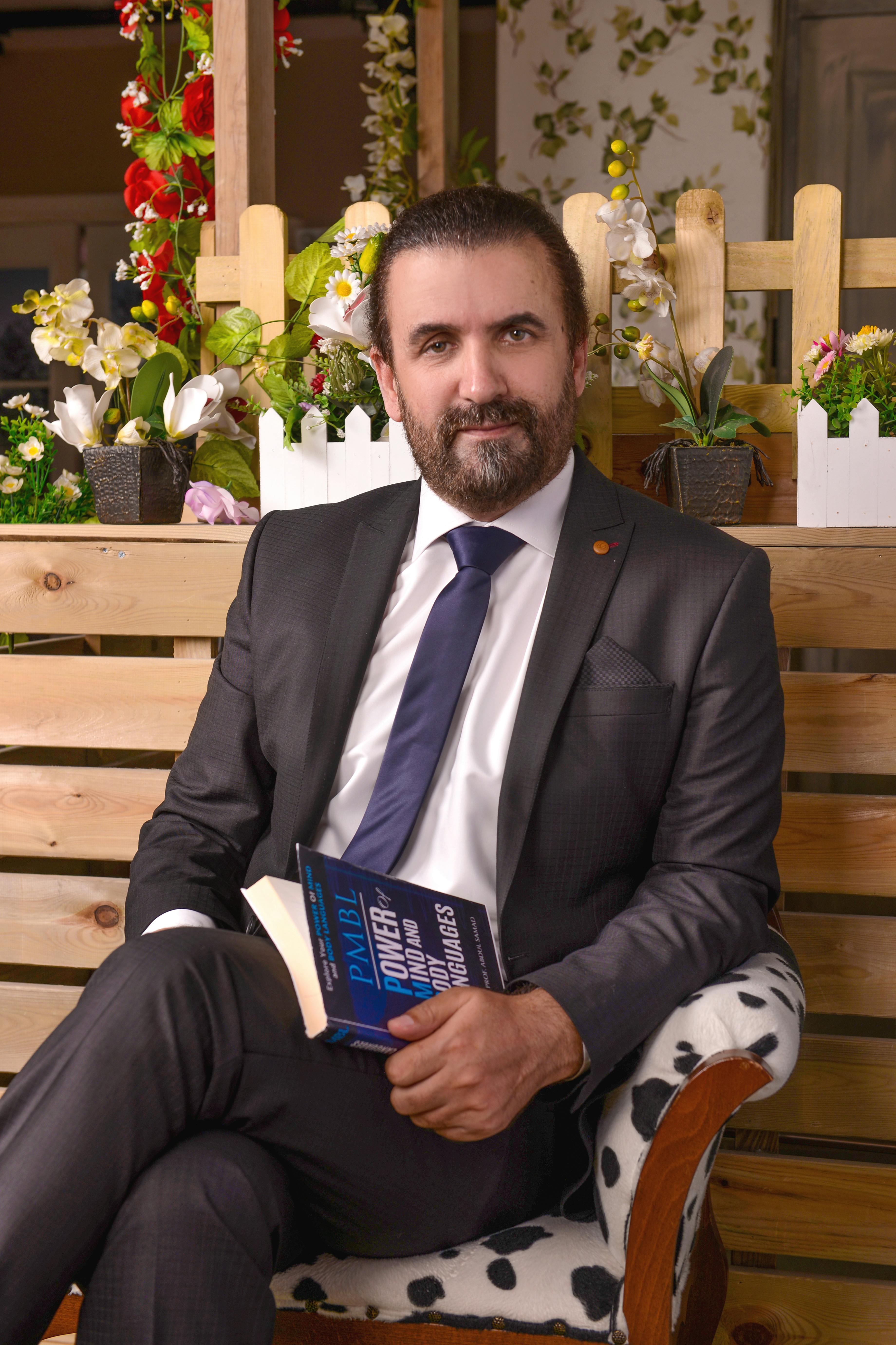 Confident businessman sitting with a book, surrounded by vibrant flowers and wooden decor.