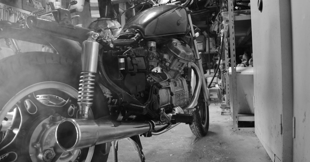 A cruiser motorcycle sits in a crowded workshop with its large exhaust and rear wheel in view
