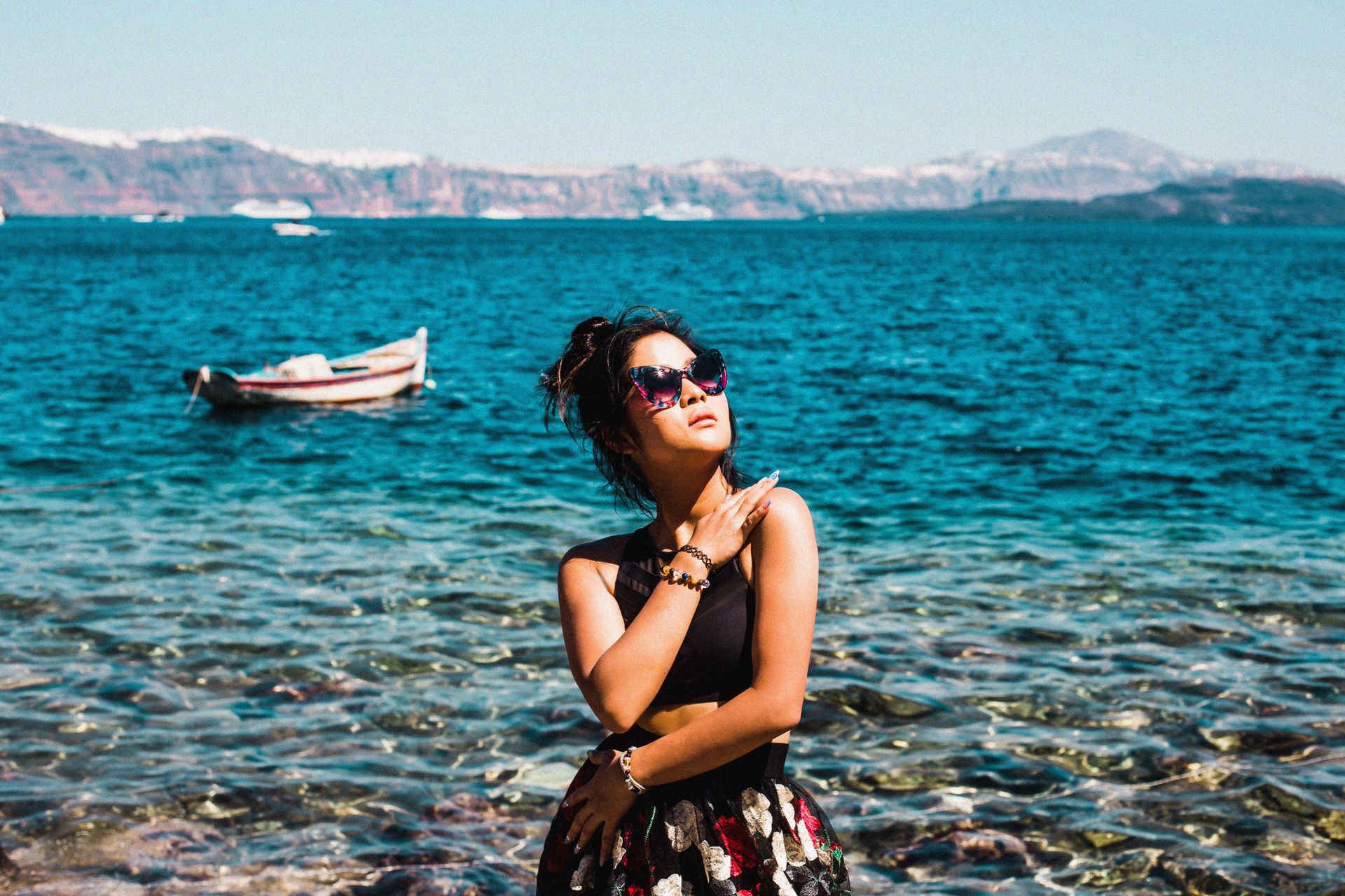 Stylish woman poses by the sparkling blue waters of a Greek beach, exuding summer vibes.