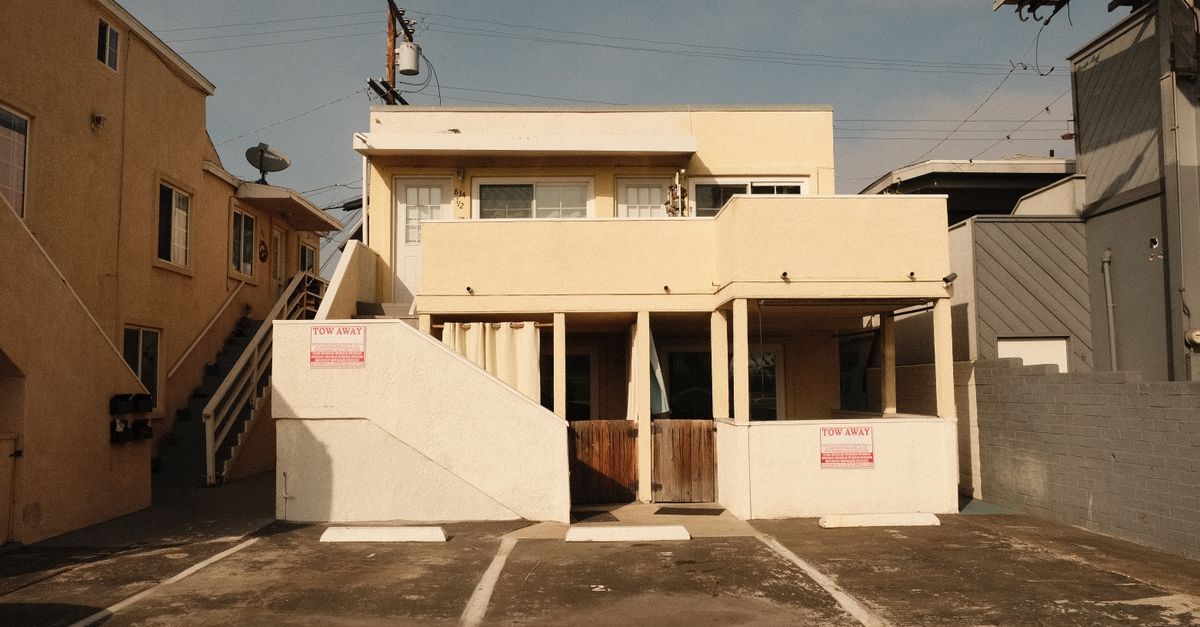A two-story beige concrete building with a flat roof showing a residential architecture style, captured in an empty driveway.