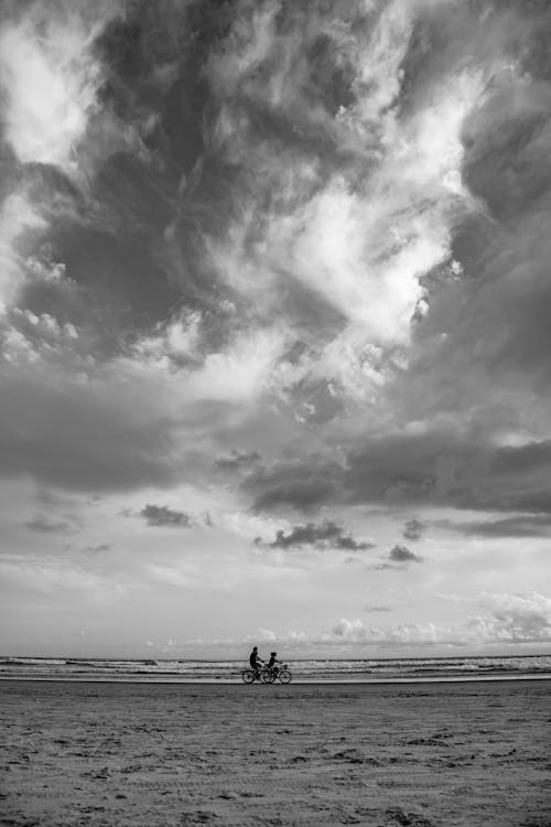 Grayscale Photo of Two People Cycling on the Beach