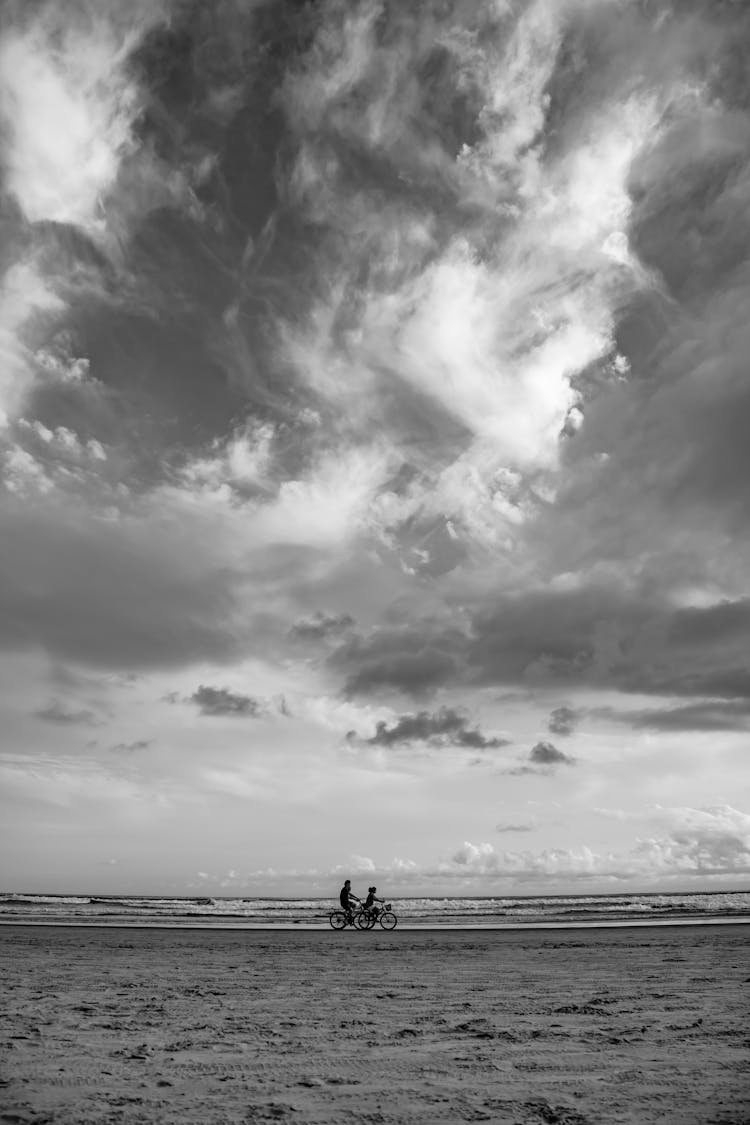 Grayscale Photo Of Two People Cycling On The Beach