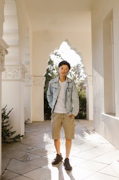 A trendy young adult stands confidently in a sunlit architectural hallway wearing casual attire.