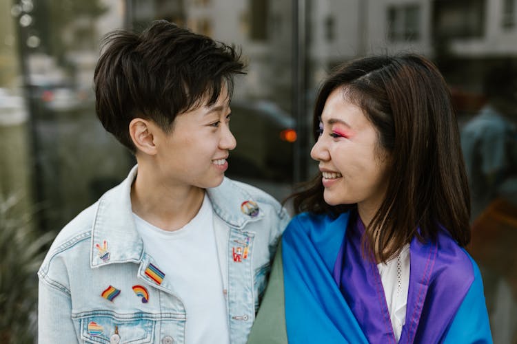 Lesbian Couple Wearing Pride Merchandise Smiling At Each Other