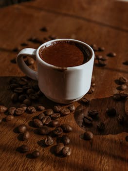 Warm coffee in a white cup with scattered beans on a wooden table.