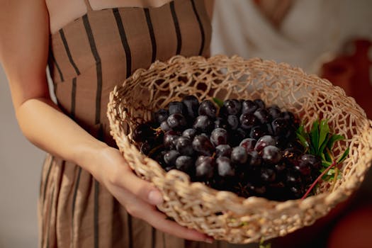 A woman in a striped dress holds a basket filled with fresh purple grapes in warm lighting.