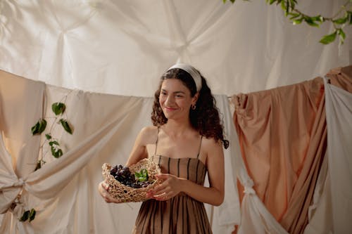 Free Smiling woman holding a basket of grapes in a warm indoor setting with draped fabrics. Stock Photo