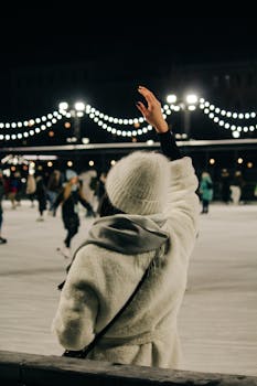 A person in winter clothes waving on an ice rink at night with festive lights.