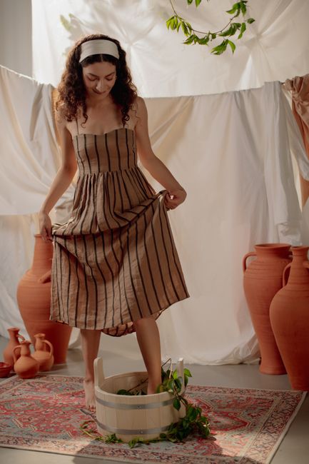 Woman in a striped dress pressing grapes barefoot surrounded by jugs in a cozy indoor setting.