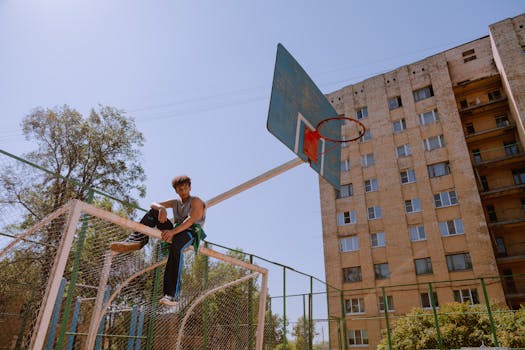 Teenager sitting on a basketball hoop, against a clear blue sky, near an urban building.