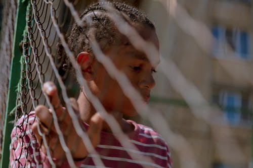 Free A Young Man in Red and White Striped Shirt Leaning on a Chain Link Fence Stock Photo