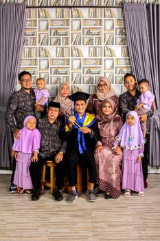 A family gathered indoors celebrating a graduation with a young adult in cap and gown. - Fotografer Sidoarjo