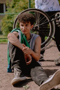 Teenager in casual clothes sitting on ground next to a bicycle in an urban area.