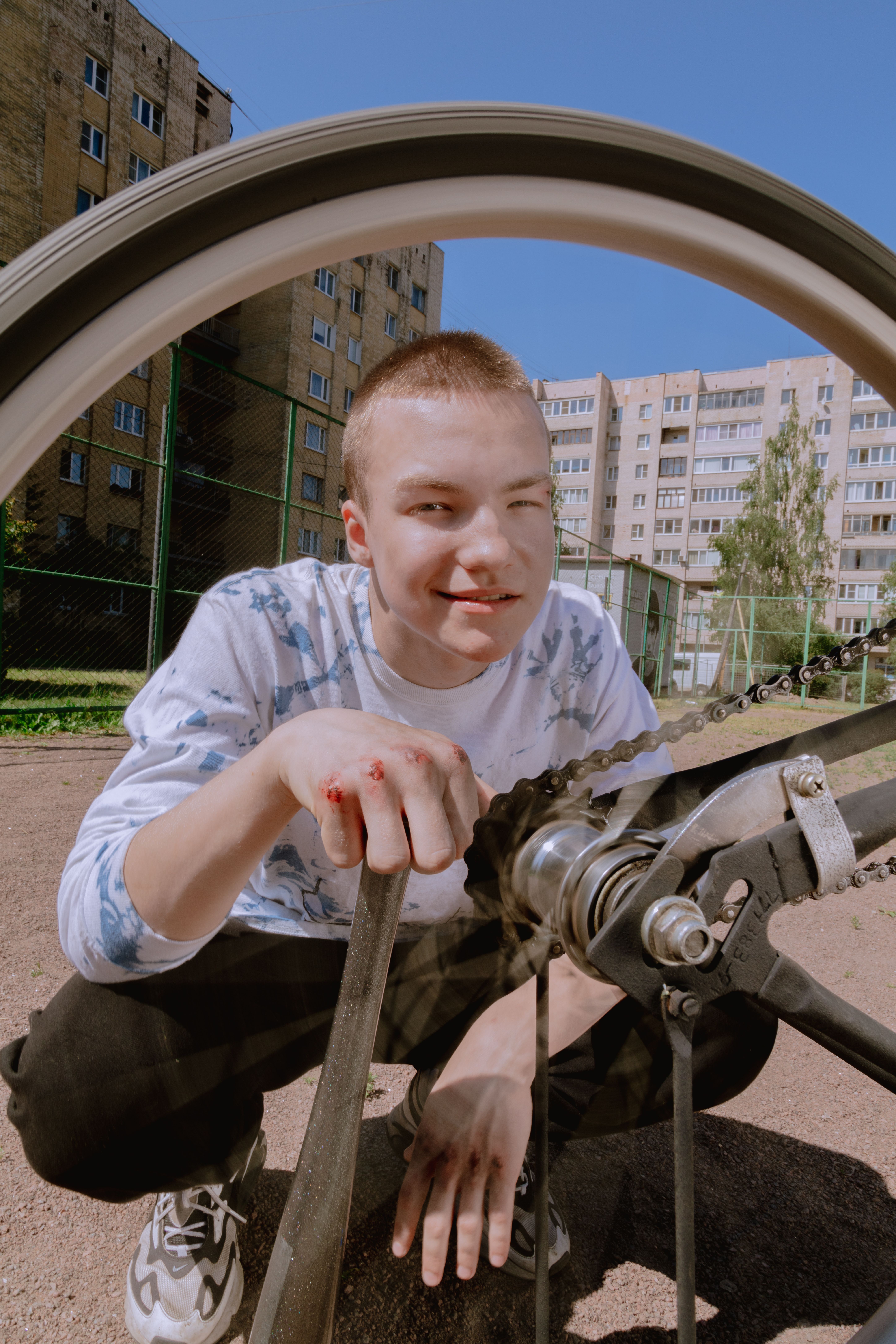 Teenager crouching to fix a bicycle in an urban setting on a sunny day.