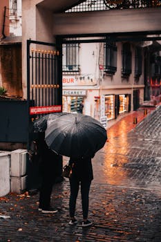 Two people with umbrellas standing by a gated entrance on a rainy street in Istanbul, Turkey.