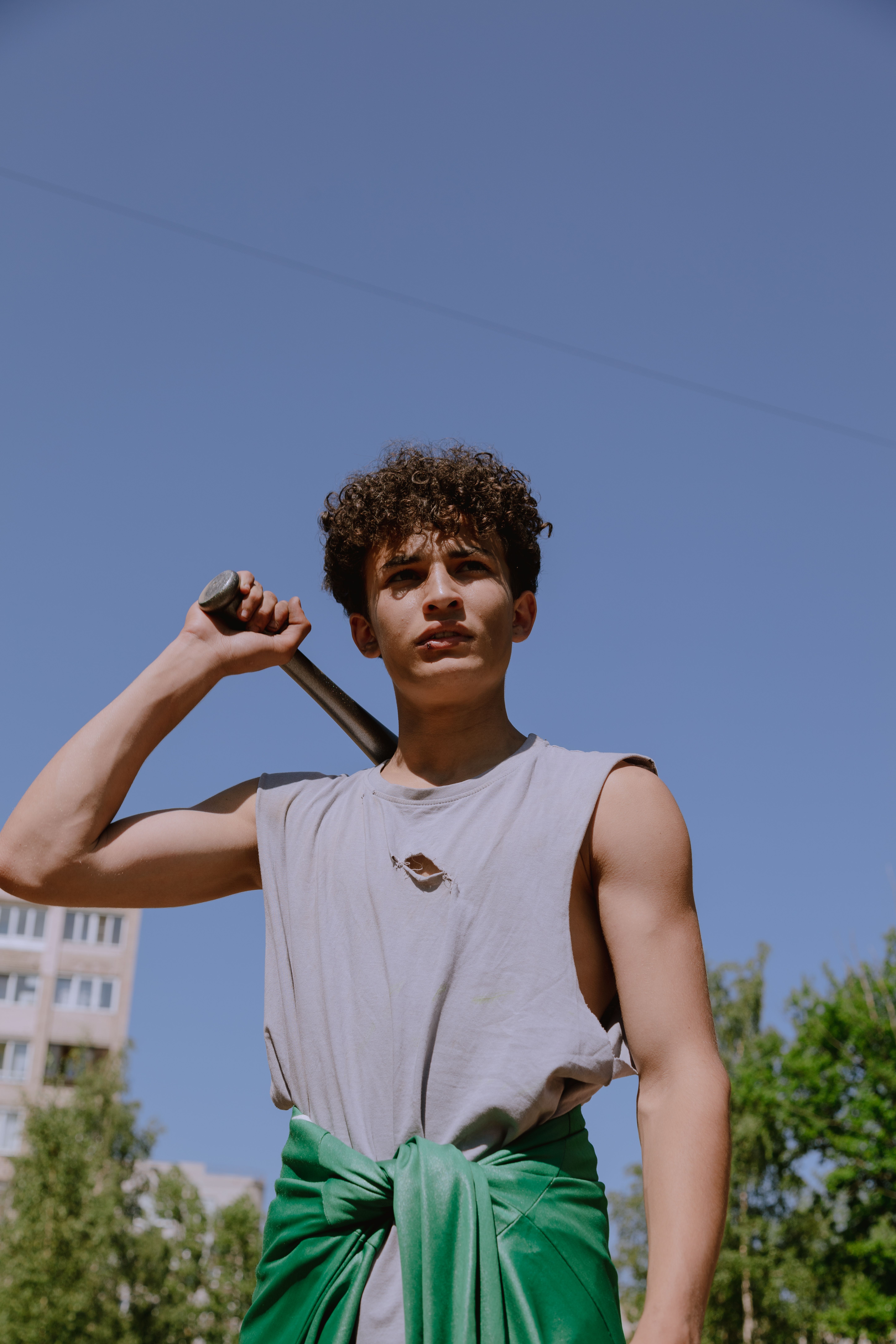 Young teenager with a bat posing confidently under a bright blue sky, wearing a sleeveless shirt.