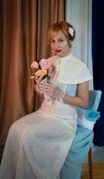 Elegant bride in white dress holding bouquet seated indoors, exuding classic charm.