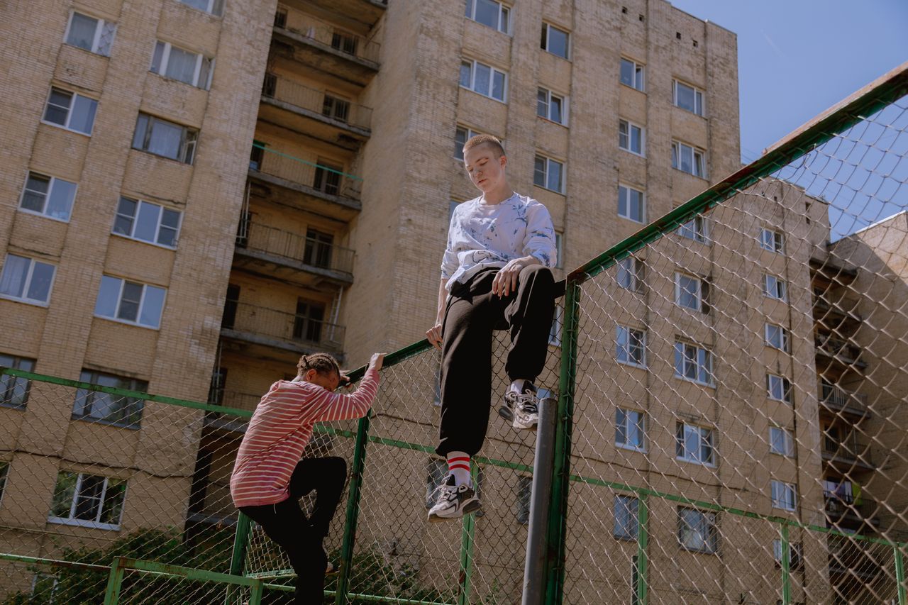 two teenagers climb a chain link fence against an urban apartment back
