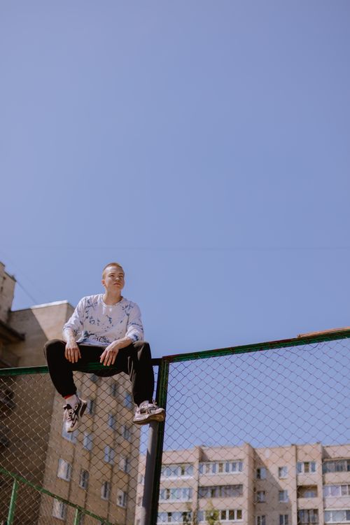 Free A stylish teenager sits on a chain link fence under a clear blue sky with urban buildings in the background. Stock Photo