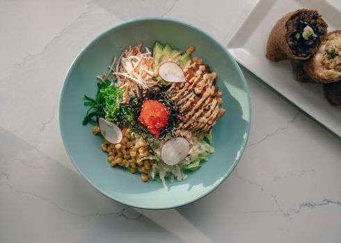Top view of a healthy poke bowl with fresh ingredients including avocado, seaweed, and corn on marble surface.
