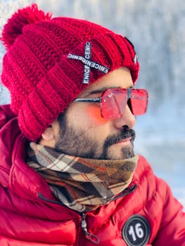 Side profile of a bearded man wearing a red knit hat and sunglasses in winter.