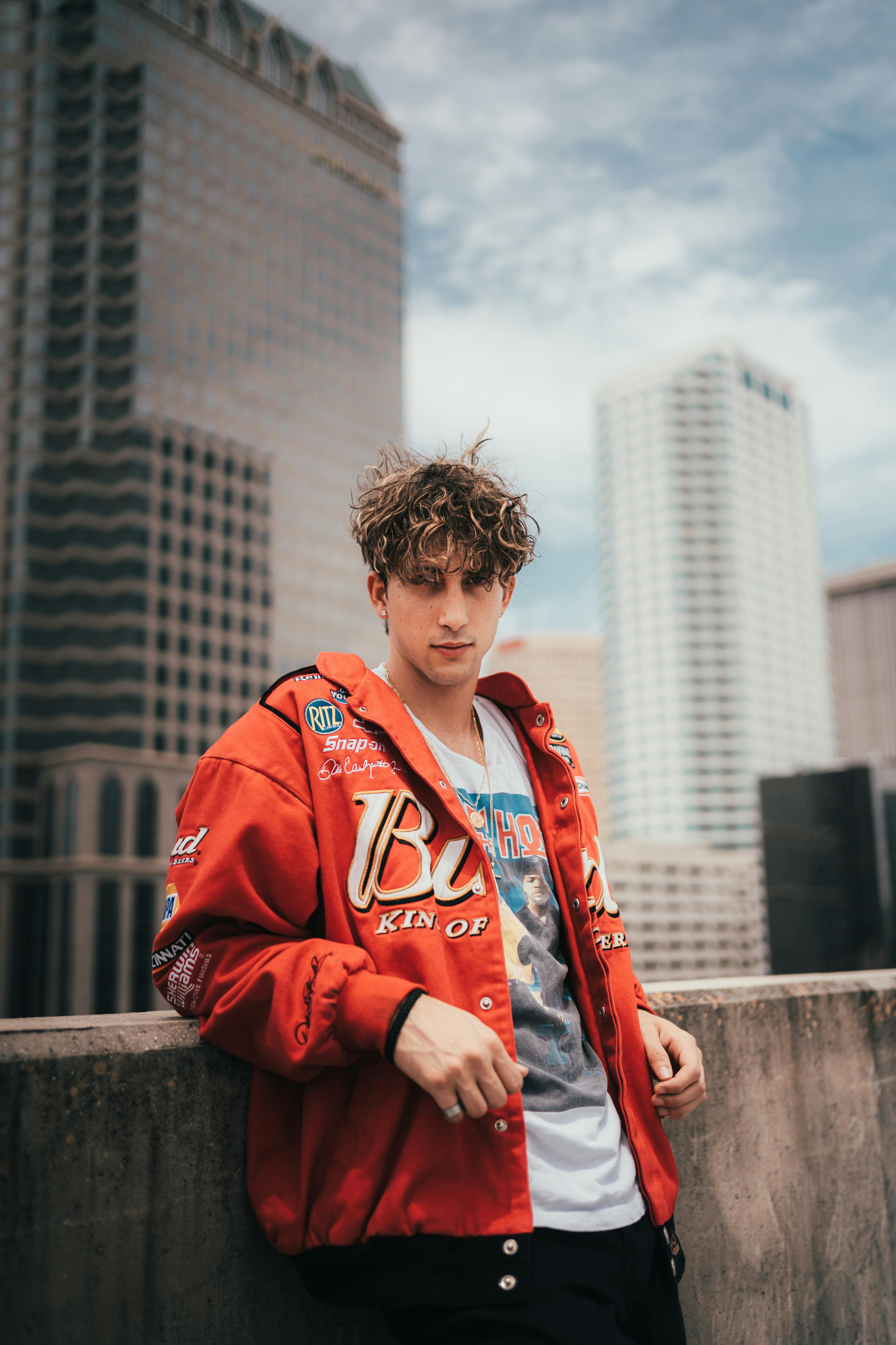 Young man in a red jacket stands confidently against a city backdrop, embodying urban street style.