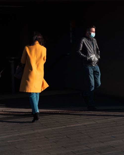 Free Urban street photo featuring a man wearing a face mask and a woman in a yellow coat walking on a sunlit pavement. Stock Photo