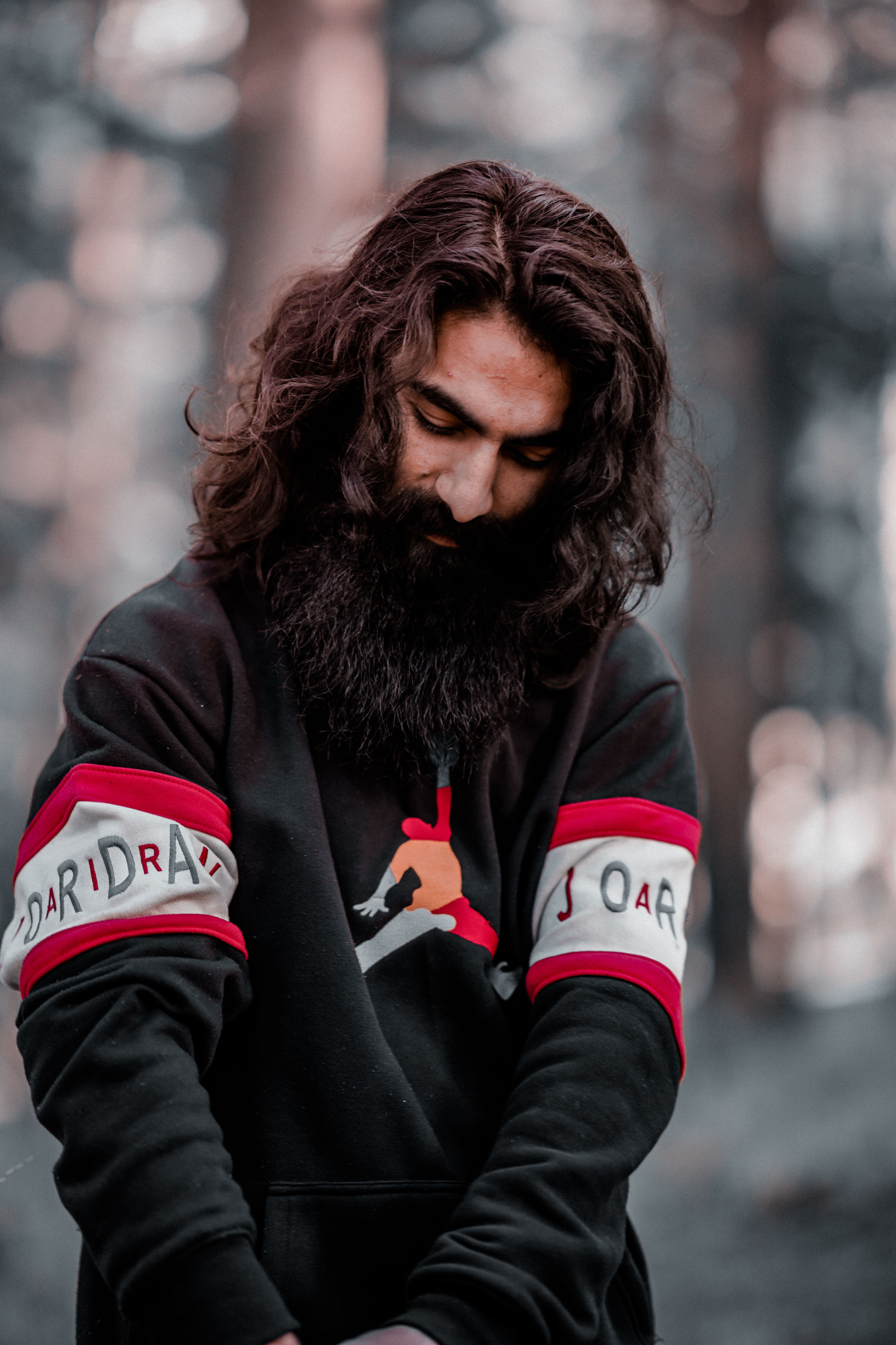 A bearded man with long hair in a sweater stands thoughtfully in a forest setting.