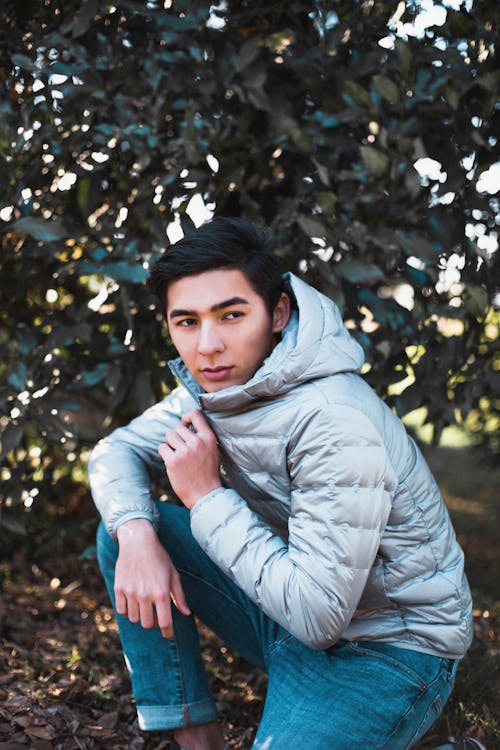 A Man in Puffer Jacket and Denim Jeans Posing Near the Green Plants