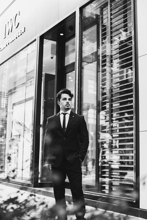 Grayscale Photo of a Man in Suit Standing Outside a Building