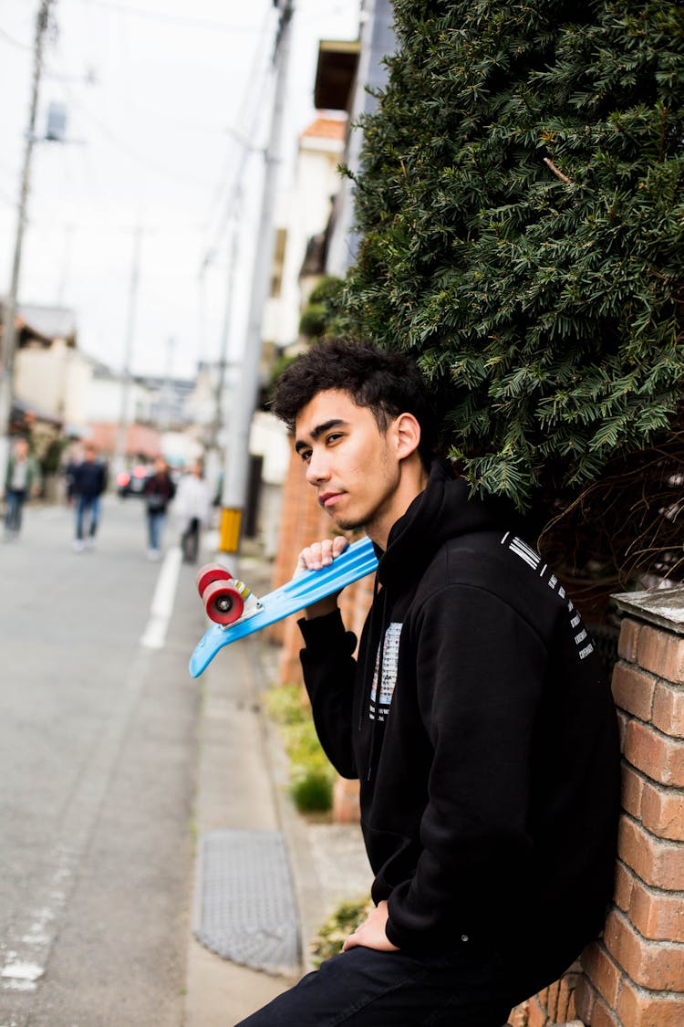 Man In A Hoodie Holding A Skateboard And Leaning On A Brick Wall