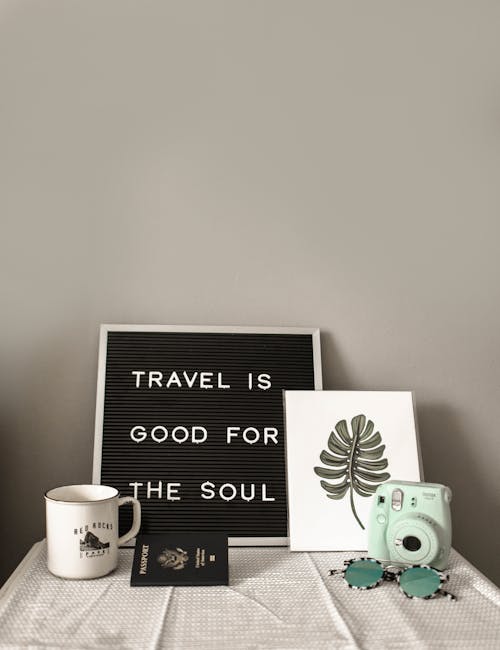 A Teal Fujifilm Instax Mini Camera laying near a white mug, palm leaf print, passport, sunglasses and a board that reads "travel is good for the soul". 