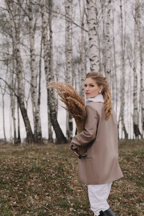 A Beautiful Woman in Brown Coat Looking Over Shoulder while Holding a Brown Dried Grass
