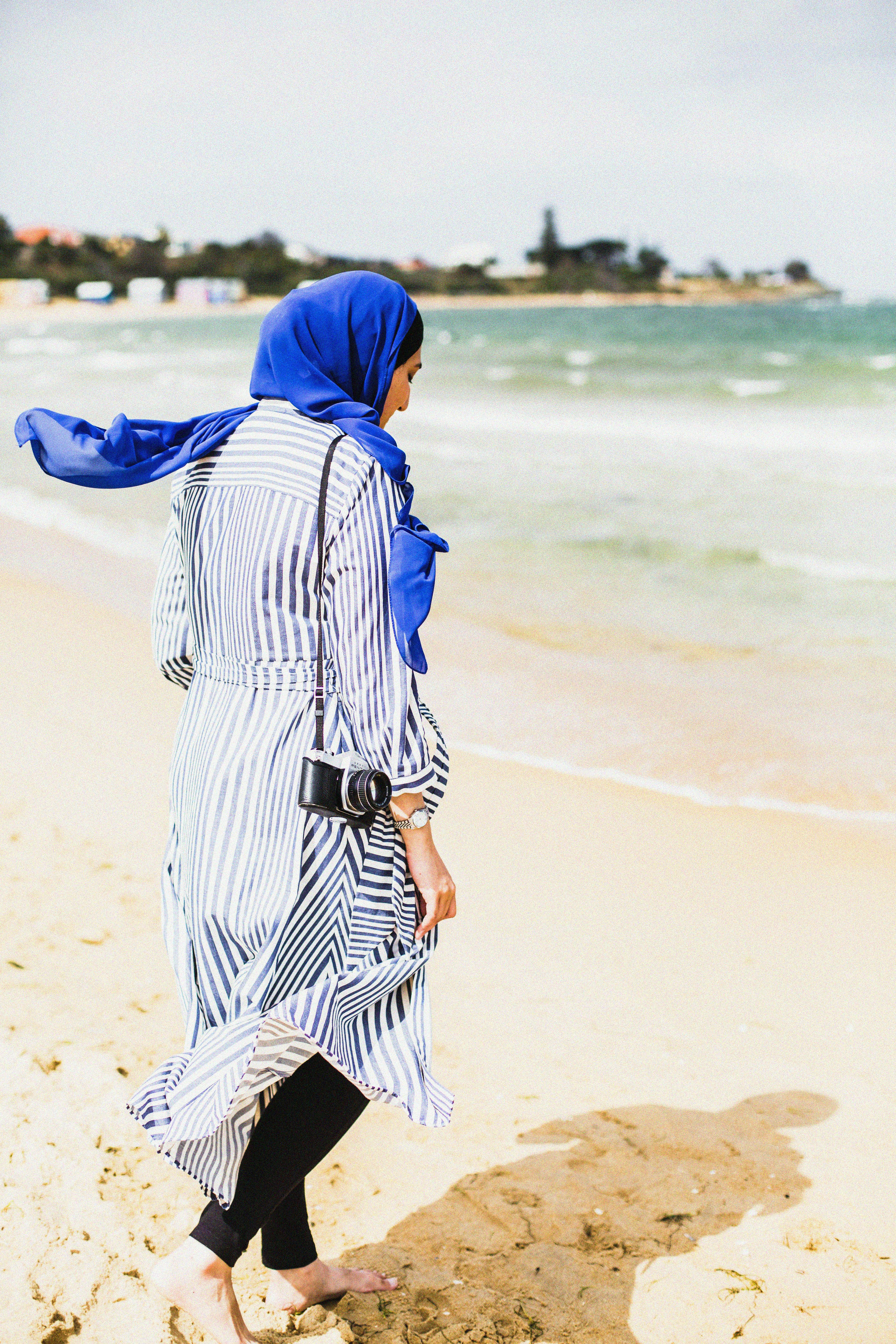 A woman in a striped dress and hijab walks barefoot on an Australian beach, carrying a camera.