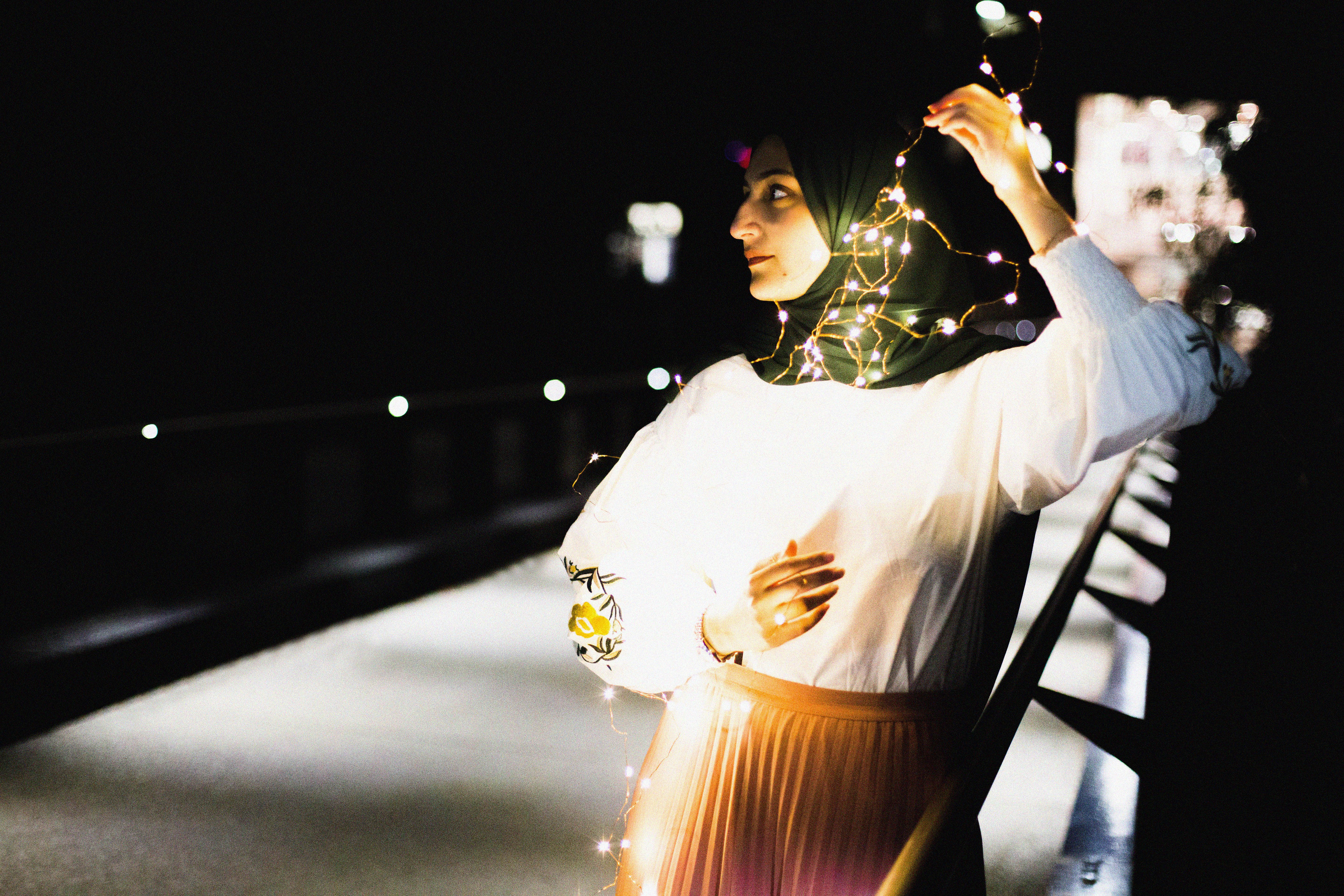 A woman elegantly poses on a bridge wrapped in fairy lights at night.
