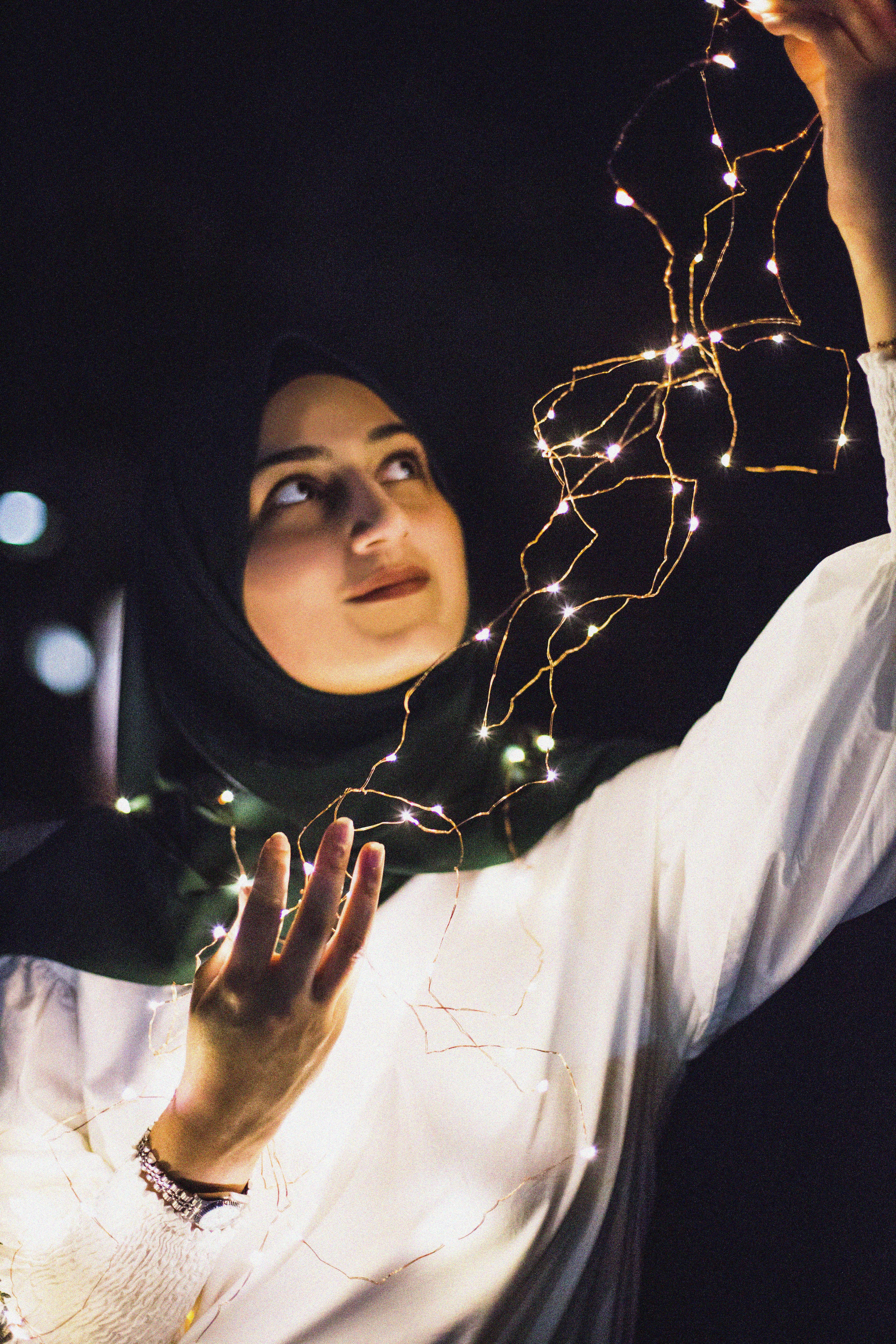 A woman in a hijab holds twinkling lights, creating a serene and magical night scene.