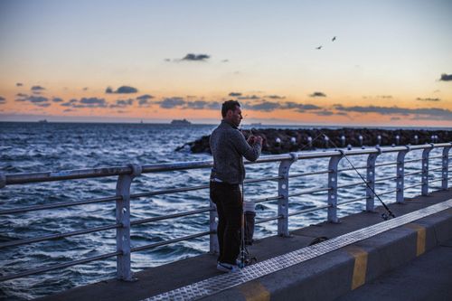 Free A man fishing on St Kilda Pier during a beautiful sunset with a view of the sea and sky. Stock Photo