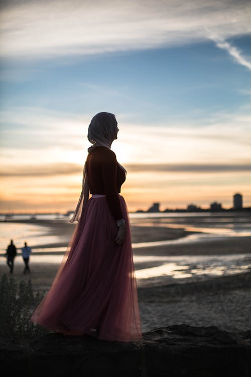 A Woman in Pink Dress with Headscarf Standing on Seashore 