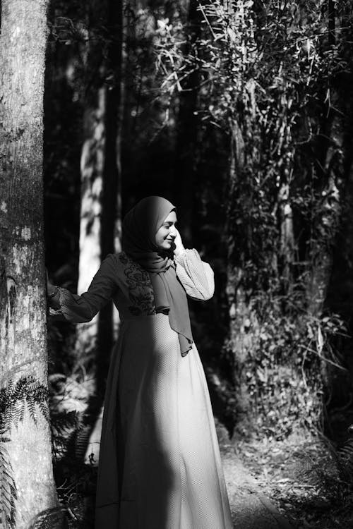 Grayscale Photo of Woman in Long Sleeve Dress and Hijab Standing Near Trees