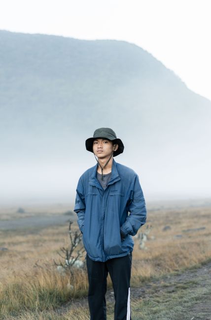 A young man in a blue jacket and bucket hat stands on a foggy mountain trail.