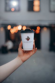 A hand holds a disposable coffee cup with a logo, against a blurred urban backdrop.
