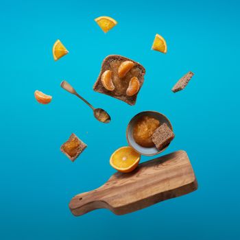 Creative still life with bread, oranges, and jam levitating against a blue background.