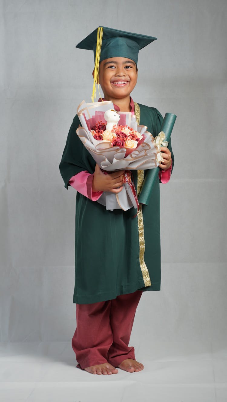 Boy Wearing A Graduation Gown With Cap Holding A Diploma And Bouquet Of Flowers